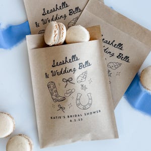 Könnte beinhalten: Geschenktüten aus Kraftpapier mit dem Text "Seashells & Wedding Bells" und einer Illustration von Cowboy-Stiefel, Hufeisen und Schleife. Die Tüten enthalten Makronen. Der Text "Katie's Bridal Shower 8.2.25" ist ebenfalls aufgedruckt.