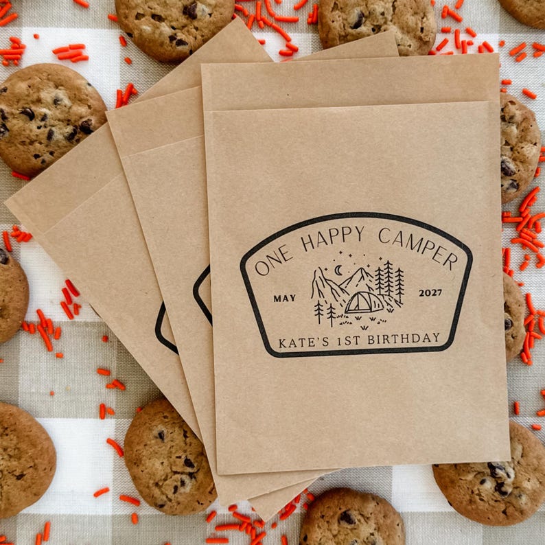 May include: A stack of light brown paper favor bags featuring a black ink stamp with a mountain and tent graphic, the text ONE HAPPY CAMPER, MAY 2027, and KATE'S 1ST BIRTHDAY. The bags are arranged on a surface with chocolate chip cookies and orange sprinkles.