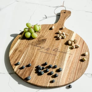 Puede incluir: Una tabla redonda de madera para servir con asa, grabada con los nombres "Lindsay & Brandon" y la fecha "September 20, 2025". La tabla está adornada con uvas verdes, arándanos y pistachos.