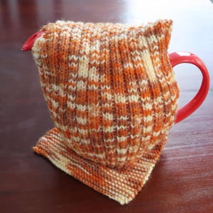 Könnte beinhalten: Eine rote Tasse mit einer gestrickten Hülle in Orange-, Braun- und Cremetönen. Die Hülle bedeckt die Tasse und hat einen passenden Untersetzer. Die Hülle ist aus einem strukturierten, gemusterten Strick gefertigt.