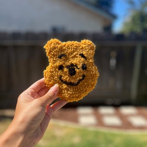 May include: A yellow felt bear with a black embroidered smiley face. The bear is being held in a hand.