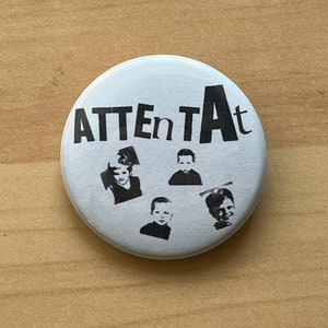 以下が含まれることがあります： 黒文字で「ATTENTAT」と書かれた白いボタンと、子供たちの顔の白黒写真が4枚ある。