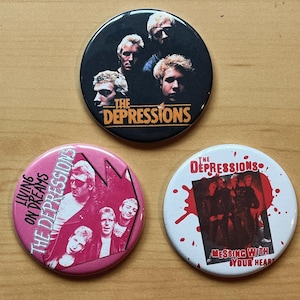 May include: Three round button pins featuring a punk rock band called "The Depressions". The first pin is black with a band photo and the text "The Depressions". The second pin is pink with a band photo and the text "Living on Dreams The Depressions". The third pin is white with a band photo and the text "The Depressions Messing With Your Heart".
