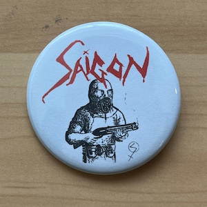 May include: A white button with a red graphic of the word "SAIGON" in a stylized font. A black and white illustration of a person wearing a gas mask and holding a shotgun is in the center of the button.