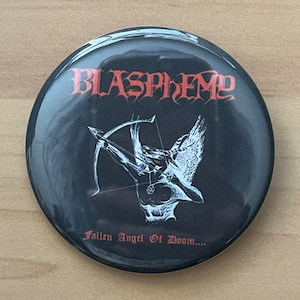 May include: Black and white button with a red band around the edge. The button features a black and white image of an angel with wings and an arrow. The text "Blasphemy" is in red at the top and "Fallen Angel Of Doom..." is in red at the bottom.