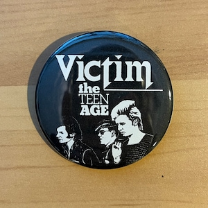 May include: Black and white button featuring a band called "Victim the Teen Age" with a photo of three people.