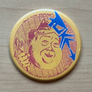 May include: A yellow button with a cartoon illustration of a person wearing a hat and holding a spoon. The text "Back to the Stone Age" and "New Album on Chickenbrain Records" is printed around the image. The button has a blue arrow pointing down.