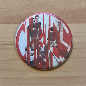 May include: A round button with a red and white design. The button features a black and white image of a band with the word "CRASS" in red letters.