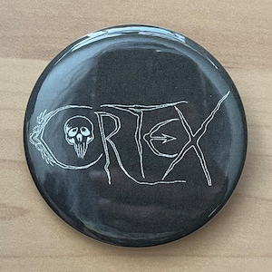 Puede incluir: Un botón negro con una calavera blanca y la palabra "CORTEX" en una fuente estilizada.
