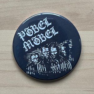 以下が含まれることがあります： ゴシック体で「Pöbel Mobel」と書かれた白黒のボタン。ボタンには、縞模様の囚人服を着た4人の白黒の画像が描かれている。