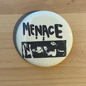 Op de afbeelding: Een witte knop met een zwart-witte afbeelding van een band genaamd "MENACE".
