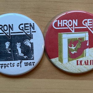 Op de afbeelding: Twee ronde witte en rode knoppen met de bandnaam "Chron Gen" erop gedrukt. De witte knop heeft een zwart-witfoto van een band met de tekst "puppets of war" eronder. De rode knop heeft een gouden en rode afbeelding van een open haard met de tekst "Reality" eronder.