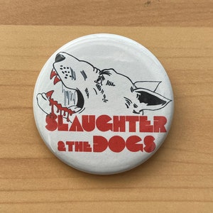 Könnte beinhalten: Ein weißer Knopf mit einer Schwarzweißillustration eines Hundekopfes mit rotem, tropfendem Blut aus seinem Maul. Der Text "SLAUGHTER & THE DOGS" steht in roten Buchstaben unter dem Hundekopf.