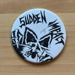 May include: A white button with a black and white illustration of a creature with claws and the text "Sudden Impact".