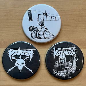 May include: Three black and white button badges featuring the word "Voivod" printed on them. The first badge depicts a black and white illustration of a tank. The second badge shows a black and white illustration of a skull with wings. The third badge displays a black and white illustration of a person wearing a helmet and holding a weapon.