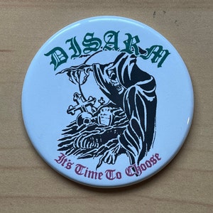 Peut inclure: Un bouton blanc avec une image en noir et blanc d'une figure capuchonnée debout au-dessus d'une pierre tombale avec une croix. Le texte "DISARM" est en lettres vertes au-dessus de la figure et "It's Time To Choose" est en lettres roses en dessous de la figure.