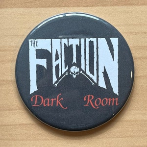 Puede incluir: Un botón blanco y negro con el texto "The Faction Dark Room" en letras blancas. La palabra "Dark" está en letras rojas. Una calavera y huesos cruzados están en el centro del botón.