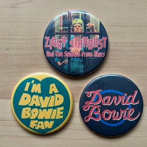 Puede incluir: Tres botones redondos con David Bowie. El primer botón es una foto de David Bowie como Ziggy Stardust con el texto "Ziggy Stardust And The Spiders From Mars". El segundo botón es verde con texto amarillo que dice "I'm A David Bowie Fan". El tercer botón es negro con texto azul y rojo que dice "David Bowie".