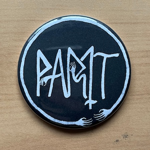 Puede incluir: Un botón negro y blanco con la palabra "PAMIT" escrita en una fuente estilizada. Las letras están formadas por manos que se extienden desde los lados del botón.