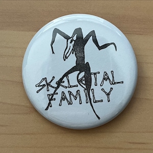 May include: White button with a black and white illustration of a skeletal figure with the text "SKELETAL FAMILY" printed below.