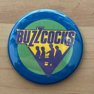 May include: A blue and yellow round button with the band name "BUZZCOCKS" in purple letters. The band's silhouette is in the center of the button.