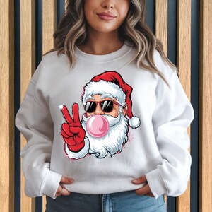 May include: White sweatshirt with a graphic of Santa Claus wearing sunglasses and blowing a bubble. Santa is giving the peace sign with his right hand.