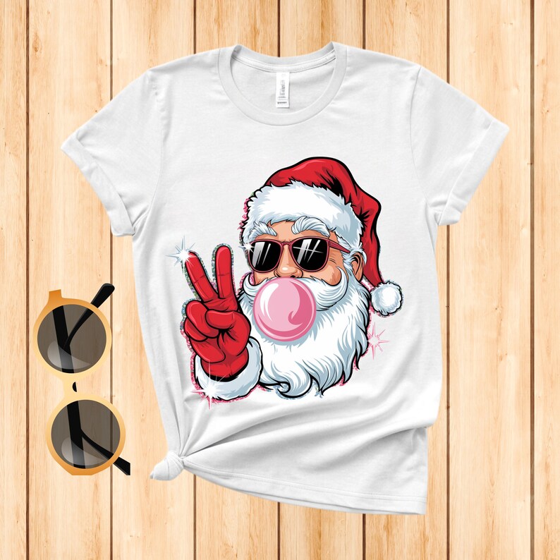 May include: White t-shirt with a cartoon image of Santa Claus wearing sunglasses and blowing a bubble with pink bubble gum. Santa is giving the peace sign with his right hand.