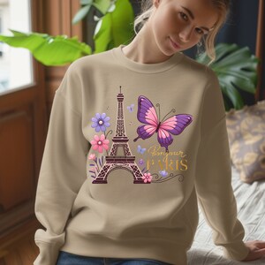 K&ouml;nnte beinhalten: Beiges Sweatshirt mit einem Aufdruck des Eiffelturms, einem lila und rosa Schmetterling und dem Text "Bonjour Paris" in Gold. Das Design umfasst lila und rosa Blumen und kleine blaue Schmetterlinge.