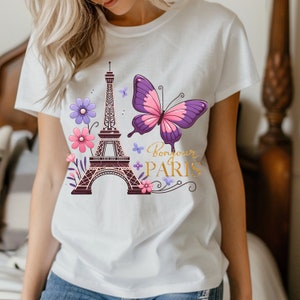 K&ouml;nnte beinhalten: Wei&szlig;es T-Shirt mit einem Design mit dem Eiffelturm, Blumen und einem Schmetterling. Die Worte "Bonjour Paris" sind in goldener Schrift geschrieben. Der Schmetterling hat rosa und lila Fl&uuml;gel.