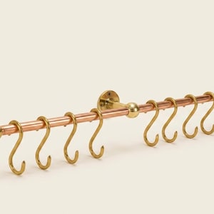 Könnte beinhalten: Eine Küchenreling aus Messing und Kupfer mit acht S-förmigen Haken. Die Reling wird mit Messingbefestigungen an der Wand befestigt. Die Kupferstange ist ca. 61 cm lang.