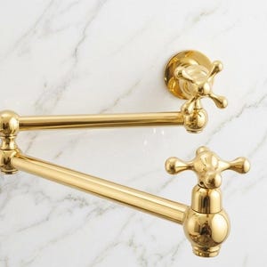 Peut inclure: Un robinet de remplissage de pot mural doré avec une conception à poignée en croix. Le robinet est doté d'un bras pivotant et est placé sur un fond de marbre blanc. Le luminaire est en laiton poli.