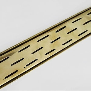 May include: A rectangular brass drain cover with a series of evenly spaced, elongated slots. The cover has a slightly aged appearance, with a warm, golden hue. The design is simple and functional, suitable for bathroom or shower applications.