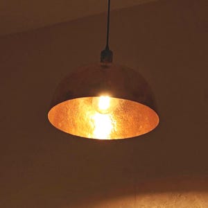 Może przedstawiać: Miedziana lampa wisząca z teksturowanym, miskowatym kloszem. Wnętrze klosza świeci ciepłym, złotym światłem z odsłoniętej żarówki. Lampa wisi na czarnym kablu.