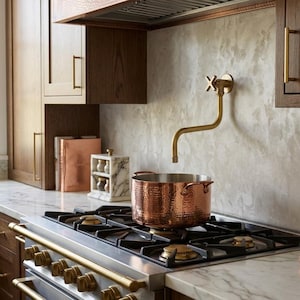 Puede incluir: Escena de cocina con una olla de cobre sobre una estufa de gas. La estufa tiene detalles en latón, y la cocina cuenta con gabinetes de madera y una encimera de mármol. Un grifo de latón está montado en la pared.