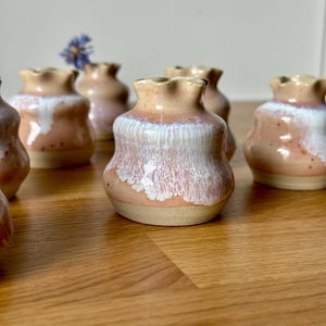 Könnte beinhalten: Eine Sammlung kleiner, handgefertigter Keramikvasen. Jede Vase hat eine einzigartige Form mit einem gerüschten Rand und einer gesprenkelten, zweifarbigen Glasur in Rosa-, Weiß- und Beigetönen. Die Vasen sind auf einer Holzoberfläche angeordnet.