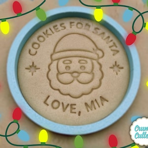 May include: A round cookie with a Santa Claus face and the text "COOKIES FOR SANTA LOVE, MIA" is inside a light blue circle. The cookie is surrounded by colorful Christmas lights and a Crumb Cutters logo.