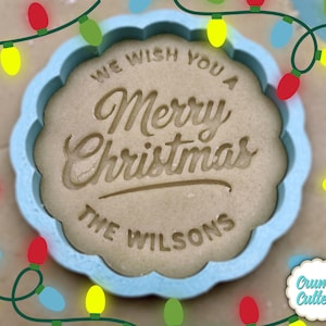 以下が含まれることがあります： 「We Wish You a Merry Christmas」と「The Wilsons」の文字が刻印された、スカラップエッジのライトブルーのクッキーカッター。背景にはカラフルなクリスマスライトがあります。