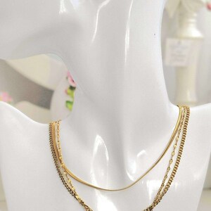 Puede incluir: Un collar de capas de oro con un pequeño colgante redondo con un cristal transparente. El collar se muestra en un busto de maniquí blanco.