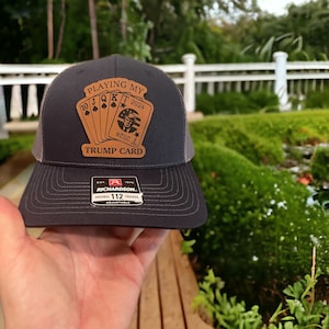 Puede incluir: Gorra de camionero gris oscuro con parte trasera de malla y visera curva. La gorra presenta un parche de cuero marrón con el texto "PLAYING MY TRUMP CARD 2024" y gráficos de naipes. La gorra es ajustable.