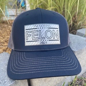 Può includere: Un cappello da camionista nero e grigio con una toppa argentata che recita "I'm Voting Felon 2024".