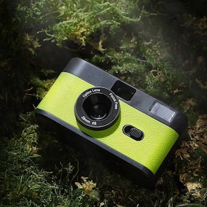 Può includere: Una macchina fotografica a pellicola da 35 mm di colore verde brillante e nero con un obiettivo da 28 mm f/8. La fotocamera ha un obiettivo a messa a fuoco libera e un flash incorporato. La fotocamera è appoggiata su un letto di muschio verde.
