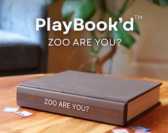 May include: A brown rectangular game box with a hinged lid, featuring the white text PlayBook'd and ZOO ARE YOU? on the cover and spine. Small square game cards with animal illustrations are scattered on the surface around the game box.
