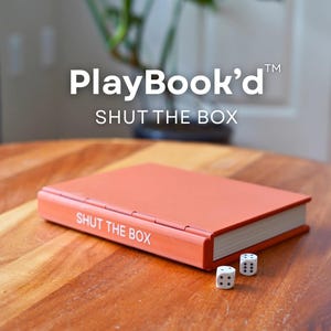 Peut inclure: Une boîte de jeu rectangulaire de couleur terre cuite portant l'inscription PlayBook'd SHUT THE BOX, dotée d'une tranche articulée et de texte blanc sur la couverture. Deux dés blancs à six faces avec des points noirs sont posés devant.