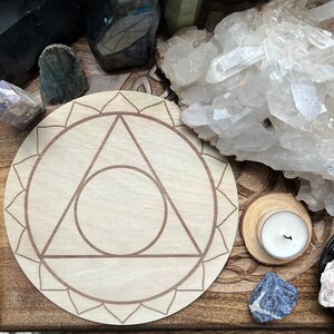 May include: A circular light-colored wooden plaque featuring a laser-engraved design of a triangle enclosed within a circle, surrounded by a petal-like border. The plaque is positioned among various raw mineral crystals and a small white tea light candle.