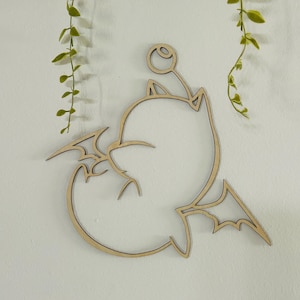 May include: Wooden wall art featuring a stylized cartoon creature with wings, ears, and a bulbous antenna. The artwork is cut from light-colored wood and mounted on a white wall, with green hanging plants in the background.