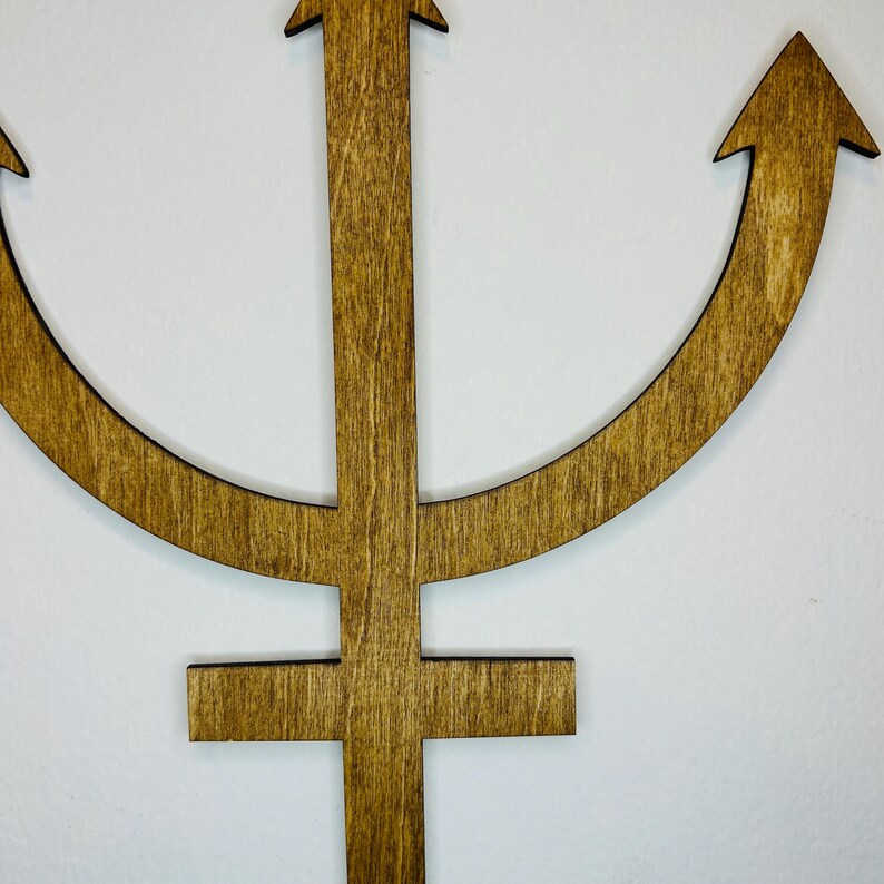 May include: A flat, brown, wooden cutout of the astrological symbol for Neptune. The design features a central vertical shaft with a horizontal crossbar at the bottom and a curved, crescent-like shape extending upwards with pointed arrow tips.