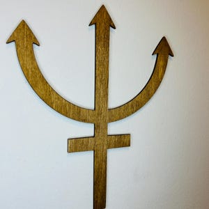 May include: A flat, brown, wooden wall decoration shaped like the astrological symbol for Neptune. The design features a central vertical shaft with a horizontal crossbar near the bottom and three upward-pointing arrows forming a crescent shape.