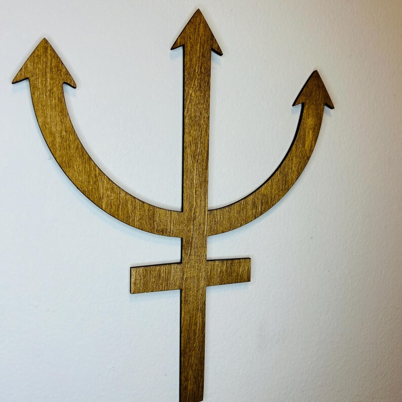 May include: A flat, brown, wooden wall decoration shaped like the astrological symbol for Neptune. The design features a central vertical shaft with a horizontal crossbar near the bottom and three upward-pointing arrows forming a crescent shape.