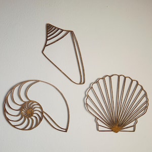 May include: Three brown wooden wall decorations shaped like seashells. One is a tall cone shell, one is a spiral nautilus shell, and one is a fan-shaped scallop shell. Each piece features a thin, open-frame design with detailed cutouts.