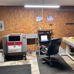 May include: A workshop space featuring a gray Boss Laser machine, a black office chair positioned at a desk with a laptop, and a wooden workbench. The room has wood-paneled walls, a fluorescent overhead light, and various tools and equipment.
